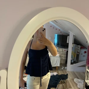 Mörkblå volangig linnetopp med knyt - Supersöt mörkblå linnetopp med volanger och knytband i halsen. Toppen har en lätt strukturerad yta och smala axelband som knyts i nacken. Perfekt för sommarens varma dagar och ger en trendig look med sina volangdetaljer.