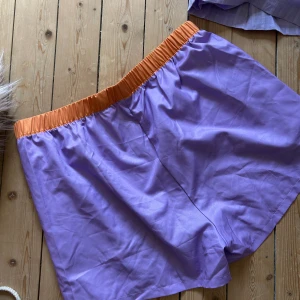 Lila shorts med orange resår - Säljer ett par loose-fit shorts i lila med bred orange resår i midjan. Perfekta för sommaren och riktigt sköna att chilla i. Materialet känns som bomull eller polyester och shortsen har en enkel, clean look utan fickor eller extra detaljer.