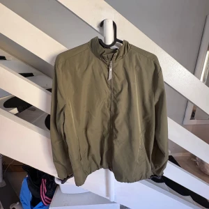 Olivgrön vindjacka från Gant 170 cm - Snygg olivgrön vindjacka från Gant med dragkedja framtill och diskret broderad logga på bröstet. Jackan har elastisk nederkant med Gant-tryck på insidan och hög krage. Perfekt för dig som gillar stilren och sportig look.