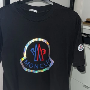 Svart Moncler t-shirt med holografiskt tryck - Svart t-shirt från Moncler med stort holografiskt logotryck i blått och rött framtill. Klassisk rund hals och korta ärmar. Liten Moncler-patch på ena ärmen. Perfekt statement-plagg för dig som gillar exklusiva streetwear-vibbar.