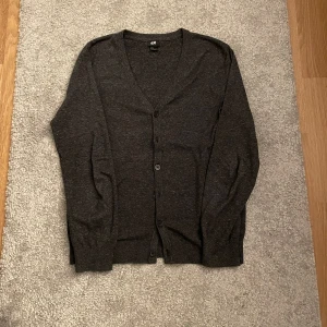Cardigan - En stilren mörkgrå kofta från H&M med v-ringning och knäppning framtill. Koftan har långa ärmar och är tillverkad i ett mjukt, tunt material som passar perfekt för lager-på-lager. Enkel och klassisk design som funkar till det mesta.