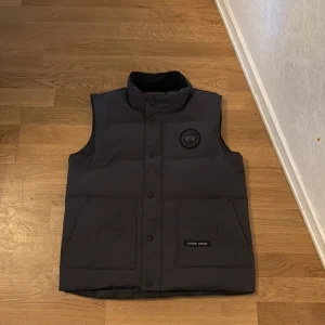 Grå dunväst från Canada Goose - Svart dunväst från Canada Goose med hög krage, tryckknappar framtill och två stora fickor. Västen har diskret logga på bröstet och klassisk quiltad design. Perfekt för lager-på-lager och håller dig varm under kyliga dagar.