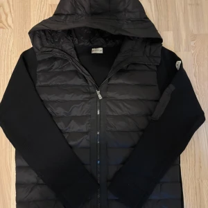 Moncler cardigan svart+NFC scan - Säljer en svart pufferjacka från Moncler med huva och ribbstickade ärmar. Jackan har dragkedja framtill och Moncler-logga på ena ärmen. Perfekt för dig som vill ha en stilren och varm jacka med snygga detaljer.