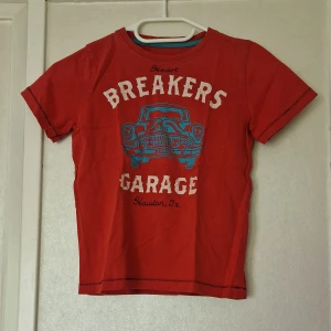 Röd t-shirt med biltryck från My wear Kids - Röd t-shirt från Mywear Kids med ett stort tryck av en blå retrobil och texten 'Heart Breakers Garage Houston, Tx' på framsidan. Rund hals och korta ärmar, tillverkad i mjuk bomull. Perfekt för dig som gillar bilar.