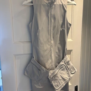 Ljusgrå ärmlös klänning från Zara - Säljer en stilren ljusgrå ärmlös klänning från Zara med hög krage och dragkedja framtill. Klänningen har två stora fickor med dragkedjor vid höften som ger en cool och modern look. 