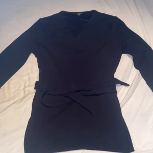 Svart omlottklänning med bälte - En stilren svart omlottklänning från Sisters Point med lång ärm och knytbälte i midjan. Klänningen har v-ringning och en enkel, elegant design som passar perfekt när du vill känna dig extra snygg. Perfekt för dig som gillar klassiska plagg med en twist.