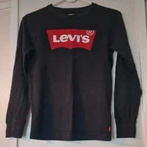 Svart långärmad Levi's tröja - Svart långärmad tröja från Levi's med klassiskt rött logotryck på bröstet. Tröjan har rund halsringning och ribbade muddar vid ärmslut.. Tillverkad i mjuk bomull som är skön mot huden. Perfekt för en avslappnad och stilren look. 