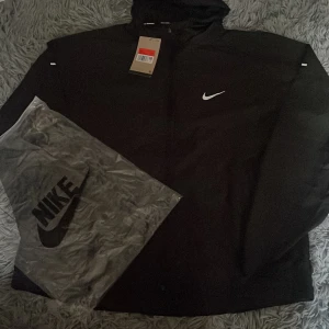 Nike miller - Svart vindjacka från Nike med vit logga på bröstet och reflexdetaljer på ärmen. Jackan har huva och dragkedja framtill. Tillverkad i lätt polyester, perfekt för löpning eller utomhusaktiviteter. Snygg och sportig design med clean look.
