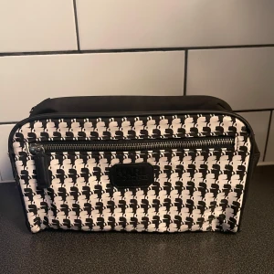 Svartvit necessär från Karl Lagerfeld - Necessär från Karl Lagerfeld med svartvitt mönster av Karl-silhuetter. Väskan har en rektangulär form, dragkedja upptill och ett extra fack med dragkedja framtill. Tillverkad i syntetmaterial och har en stilren logga på framsidan.