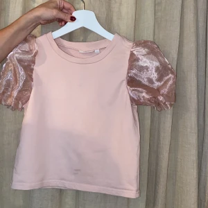 Rosa topp med puffärmar från Kappahl - Supersöt ljusrosa topp från Kappahl med glansiga puffärmar i transparent material. Kroppen är i mjuk bomull och ärmarna ger en extra touch med sitt skimrande tyg. Perfekt för dig som vill sticka ut lite extra med en trendig och lekfull look.