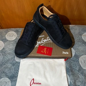 Christian Louboutin svarta sneakers  - Snygga svarta sneakers från Christian Louboutin i mjuk mocka med klassisk röd sula. Skorna har rund tå, svarta skosnören och diskret broderad logga på hälen. Perfekta för dig som vill ha en lyxig och stilren look med en ikonisk touch.