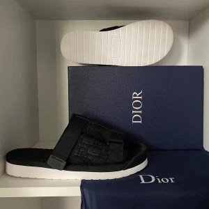 Svarta sandaler från Dior - Säljer ett par svarta Dior alpha slides med breda remmar och justerbart spänne. Ovandelen har diskret Dior-mönster och sulan är vit med tydlig Dior-logga undertill. Snygg och modern design, perfekt för sommaren. Skriv vid frågor!!!