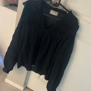 Svart blus med volang från Neo Noir - Säljer en svart blus från Neo Noir med volangdetaljer och v-ringning. Blusen har långa ärmar med lätt puff och dekorativa broderade partier. Tillverkad i ett tunt, luftigt material som ger en flowy känsla. Det är ett matchande sett med en kjol till. 