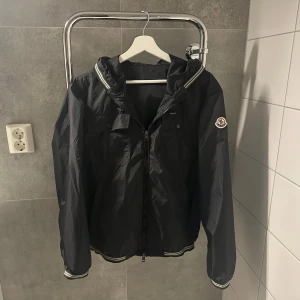 Moncler Jeanclaude Jacka - Svart vindjacka från Moncler med huva och dragkedja framtill. Jackan har två bröstfickor med knappar, Moncler-logga på ärmen och sportiga ränder vid muddar och huva. Perfekt för dig som vill ha en clean och stilren look. Mycket bra skick. Storlek 2 vilket motsvarar runt M-L