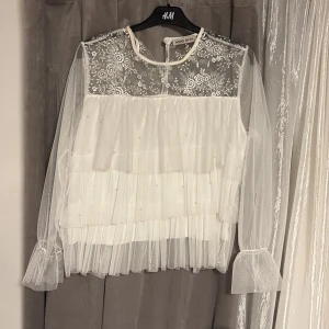 Vit blus med spets och volanger  - Superfin vit blus med transparent mesh, broderade blommor upptill och flera lager volanger. Långa ärmar i tunn mesh med lätt volang vid ärmslut.