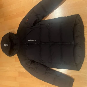 Dunjacka, Sail racing  - Säljer en svart dunjacka från Sail racing. Jackan har en hög krage och huva för extrem värme den har två bröst fickor samt två nere med dragkedja framtill och har en stilren design, snabb affär uppskattas.