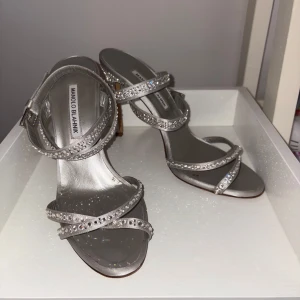 Manolo Blahnik silverglittriga sandaler - Snygga silverfärgade sandaler från Manolo Blahnik med smala remmar och glittrande stenar över hela skon och klacken. Klackarna är höga och smala, och remmarna har spänne vid ankeln. Perfekta för dig som vill sticka ut med extra bling.