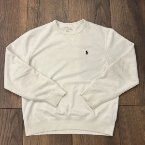 Vit sweatshirt från Polo Ralph Lauren - Klassisk vit sweatshirt från Polo Ralph Lauren med rund hals och broderad svart logga på bröstet. Tröjan har långa ärmar och ribbade muddar vid ärmslut och nederkant. Perfekt för en clean och stilren look.