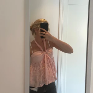 Ljusrosa halterneck linnetopp - Supersöt ljusrosa linnetopp med halterneck och knyt fram. Toppen har en flowy, luftig passform och smockad rygg. Perfekt för varma dagar och ger en romantisk vibe. Materialet känns tunt och lätt, och toppen är ärmlös.