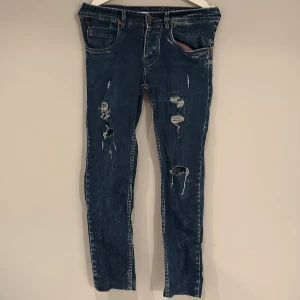Blå slitna jeans med hål - Säljer ett par blå jeans med slitningar och hål på benen för en riktigt edgy look. Jeansen har klassisk femficksdesign och raka ben. Perfekta för dig som gillar streetstyle och vill sticka ut lite extra.