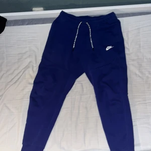 Blå mjukisbyxor från Nike med snörning - Snygga blå mjukisbyxor från Nike med vit logga på benet och dragsko i midjan. Byxorna har smal passform, muddar vid bensluten och dragkedjeficka bak. Perfekta för chill eller träning.
