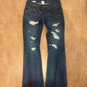 True Religion distressed flared jeans blå - Snygga blå jeans från True Religion med slitna detaljer och flared passform. Klassiska kontrastsömmar, femficksmodell och ikoniska bakfickor med lock och knapp. Jeansen har låg midja och är tillverkade i kraftigt denimtyg.