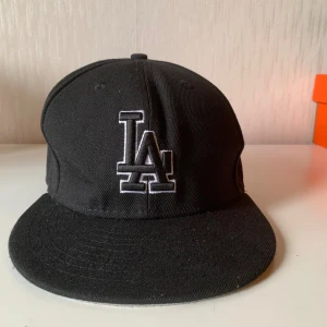 Svart LA-keps från New Era 59FIFTY - Säljer en svart New Era 59FIFTY keps med LA-logga broderad framtill i vitt. Klassisk basebollmodell med rak skärm och MLB-logga baktill. Kepsen är gjord i polyester och har en stilren look som passar till streetwear.
