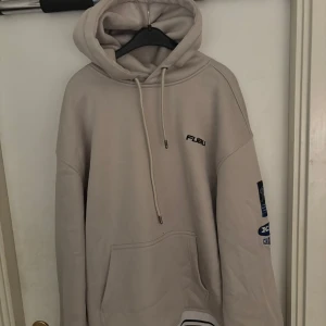 Beige FUBU hoodie med tryck och patchar - Säljer en beige hoodie från FUBU med coola tryck och patchar på både bröstet, ryggen och ena ärmen. Tröjan har huva med snörning, känguruficka och sportig vibe. Materialet är polyester och modellen är loose fit. Perfekt för dig som gillar streetwear. Funkar även för L!!