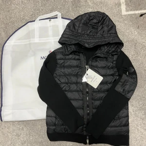 Svart Moncler dunjacka med huva - Svart dunjacka från Moncler med quiltad front och huva. Jackan har stickade ärmar och sidor, dragkedja framtill och en ficka med dragkedja på ärmen. Klassisk Moncler-logga på insidan och coolt serietryck. Perfekt för dig som vill ha en stilren och sportig look.