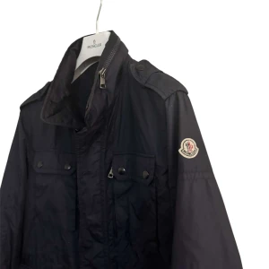 Svart vindjacka från Moncler - Svart vindjacka från Moncler med klassisk logga på ärmen, hög krage och axelklaffar. Jackan har två bröstfickor med knappar och dragkedja framtill. Tillverkad i lätt polyester, perfekt för blåsiga dagar.