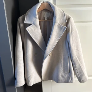 Beige skepparkavaj från H&M - Stilren beige skepparkavaj från H&M med breda slag, dubbelknäppning och raka fickor. Jackan har en klassisk, tidlös look och är fodrad för extra komfort. Perfekt för dig som vill ha en clean och snygg stil.