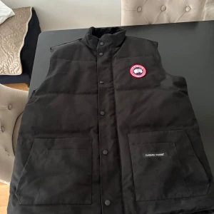 Svart dunväst från Canada Goose XXL - Svart dunväst från Canada Goose i storlek XXL. Västen har hög krage, två stora framfickor med logotyp, tryckknappar och det klassiska röda och vita Canada Goose-märket på bröstet. Perfekt för kalla dagar och har en stilren. Storleken är då XXL men sitter mer som XL/L och är väldigt varm och skön passar perfekt för höst och en bit på vinter. tyvär finns inget kvitto!  Säljs för 400