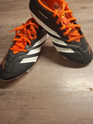 Adidas Predator fotbollsskor svart/orange - Säljer ett par Adidas Predator fotbollsskor i svart med vita detaljer och orange snörning. Skorna har klassiska tre ränder på sidorna, orange insida och dobbar för gräsplan. Ovandelen är i syntetmaterial och designen är riktigt sportig. Minimalt smutsiga men går ju självklart bort om man tvättar bort! 