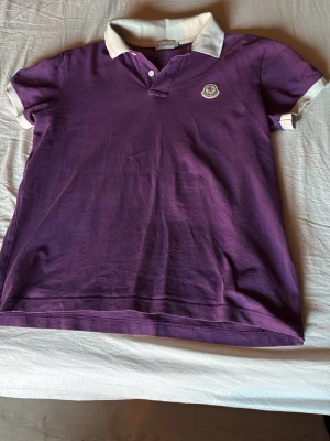 Moncler pikétröja med vit krage S/M - Säljer en lila pikétröja från Moncler med vit krage och vita ärmslut. Tröjan har knappslå framtill och Moncler-logga på bröstet. Klassisk passform och mjukt bomullsmaterial. Moncler-broderi på baksidan av kragen för extra detalj. Obs! Sitter som S 