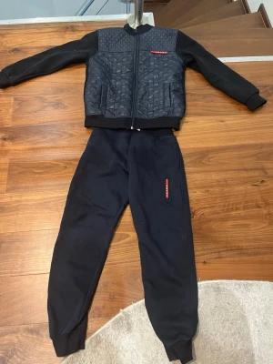 Svart quiltad väst från Prada - Säljer en svart quiltad väst från Prada med cool struktur på framsidan och diskret röd logga. Västen har dragkedja framtill och två fickor. Materialet är 85% bomull och 15% polyester, vilket gör den både mjuk och bekväm.