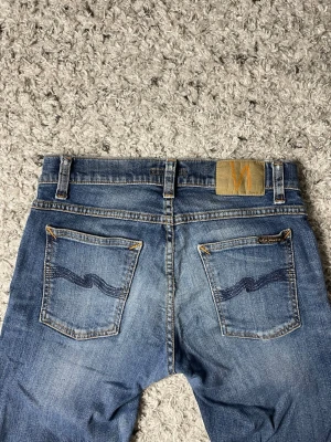Blå jeans från Nudie Jeans - Säljer ett par klassiska blå jeans från Nudie Jeans med snygga slitningar och kontrastsömmar. Jeansen har fem fickor, orange patch bak och den ikoniska vågstickningen på bakfickorna. Perfekta för dig som gillar en smal passform och tidlös stil.