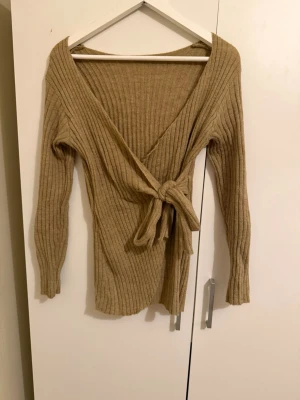 Beige ribbad omlottopp med knyt - Säljer en beige ribbad omlottopp med lång ärm och snygg knytning i midjan. Tröjan har djup v-ringning och är gjord i ett mjukt stickat material som känns både trendigt och bekvämt. Perfekt att styla med jeans eller kjol för en chill look.