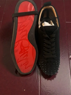 Äkta Christian Louboutin svarta sneakers med nitar med kvitto - Säljer ett par svarta sneakers från Christian Louboutin i mocka med ikonisk röd sula och svarta nitar över tån. Skorna har snörning och en rund tå. Kommer med originalkartong och dustbag. Perfekta för dig som vill sticka ut med en edgy och lyxig look. Kommer med Kvitto