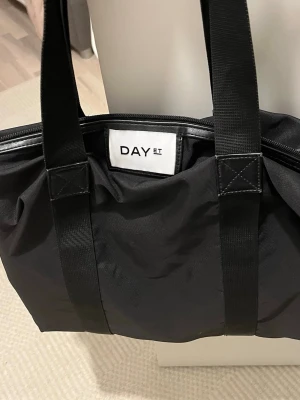 Svart axelväska från DAY ET - Stilren svart axelväska från DAY ET med rymligt huvudfack och innerficka med dragkedja. Väskan är tillverkad i slitstark polyester och har långa, breda axelband för enkel bäring. Perfekt för dig som vill ha plats för allt du behöver under dagen.