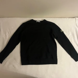 Svart sweatshirt från Stone Island - Svart sweatshirt från Stone Island med klassisk rund hals och lång ärm. Ikonisk logotyp-patch på vänster ärm. Tröjan är i bomull och har en clean, stilren look som passar perfekt till streetwear. Enkel och snygg design utan mönster. (Använd fåtal gånger) 