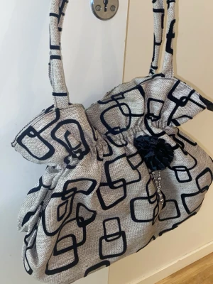 Beige handväska med svart mönster - Säljer en beige handväska med coolt svart grafiskt mönster och en dekorativ svart blomdetalj framtill. Väskan har två handtag och är gjord i ett tygmaterial som känns lite grovt. Perfekt för dig som vill sticka ut med en unik accessoar.