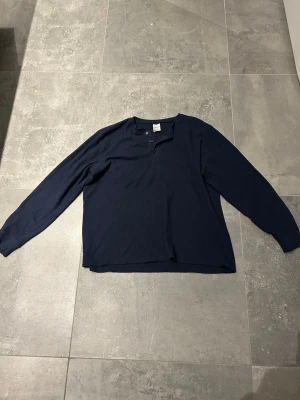 Mörkblå henleyshirt med knappar - En enkel och stilren mörkblå henleyshirt med två knappar vid halsen. Tröjan har en rak passform och är gjord i mjuk bomull, perfekt för lager-på-lager under kyligare dagar.