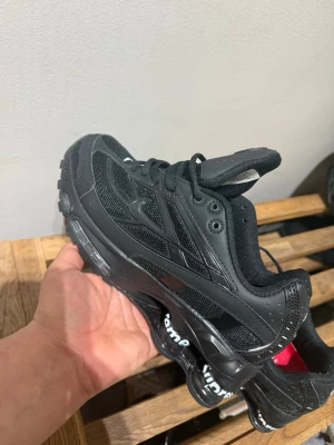 Nike x Supreme svarta sneakers - Nike x Supreme sneakers i helsvart med Supreme-logga på sulan och röd innersula. Skorna har mesh och syntetpaneler, snörning och tydliga Nike-detaljer. Grymt chunky sula och sportig vibe, perfekt för dig som gillar streetwear.