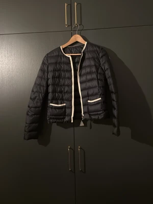 Svart Moncler pufferjacka med vita detaljer - Säljer en svart cardigan aktig jacka från Moncler med vita kontrastkanter och två fickor framtill. Jackan har rund halsringning, quiltad design och dragkedja. Perfekt för dig som vill ha en lätt och snygg jacka till vardags. Klassisk Moncler-logga på ärmen. Jackan är knapp använd och säljs inte längre så passa på!! Ifall ni vill se bevis på att den är äkta är det bara och skicka ett meddelande!