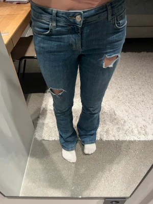 Blå bootcut jeans från Zara - Säljer ett par blå bootcut jeans från Zara med slitningar och hål på båda knäna. Jeansen har låg midja och klassisk femficksdesign. Materialet är jeans i bomull och de har en snygg, lite utsvängd passform nertill. Använd helst inte köp direkt! Skriv för mer info 😊