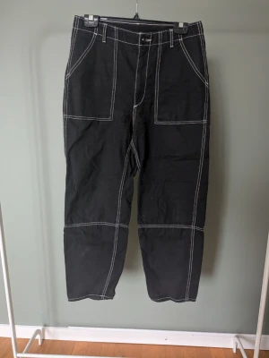 Svarta "snickar" jeans med kontrastsömmar - Svarta jeans med vita kontrastsömmar och stora fickor fram och bak. Byxorna har raka ben och bälteshällor i midjan. Materialet känns slitstarkt och de har en cool, loose passform som ger en avslappnad vibe.