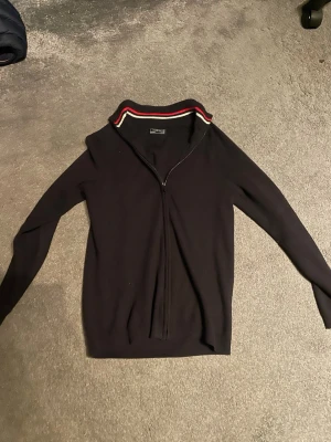 Svart kofta med dragkedja  - Säljer en svart kofta med dragkedja och ribbad krage med röda och vita ränder. Tröjan är långärmad och har en clean, stilren look som passar perfekt till jeans eller chinos.