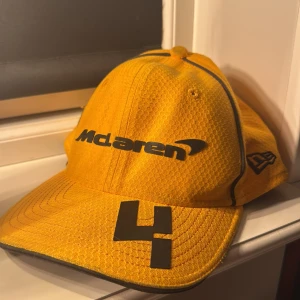 Gul McLaren keps med svart logga - Säljer en gul McLaren keps med svart logga och siffran 4 framtill. Kepsen har sportig design med mesh-liknande material och svarta detaljer längs sömmarna. Perfekt för dig som gillar racing och vill sticka ut med en färgstark accessoar.