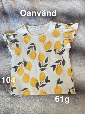 Vit t-shirt med citronmönster Lindex - Superfin vit t-shirt från Lindex med färgglada citroner och gröna blad över hela. T-shirten har korta ärmar med volangdetalj. Perfekt för dig som gillar lekfulla prints och somriga vibes!🌸 Ny utan lapp ( ej upptvättad) storlek 104 