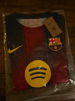 FC Barcelona Nike matchtröja - Säljer en officiell FC Barcelona matchtröja från Nike med klassiska blå och röda ränder, gult Nike-märke och klubbmärke på bröstet. Framsidan har stor gul Spotify-logga och baksidan UNHCR-tryck. Tillverkad i lätt polyester, perfekt för fotboll eller samling.